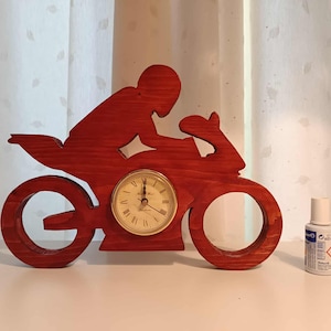 Könnte beinhalten: Eine rote Holz-Motorrad-Uhr mit einer Fahrer-Silhouette. Das Zifferblatt ist goldfarben mit schwarzen Zeigern und römischen Ziffern. Rechts von der Uhr steht eine kleine Flasche. Die Uhr ist etwa 25 cm breit.