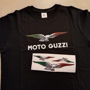 Könnte beinhalten: Schwarzes T-Shirt mit dem Moto Guzzi-Logo, einem stilisierten Adler mit grünen, weißen und orangefarbenen Akzenten. Der Text "MOTO GUZZI" ist darunter gedruckt. Ein Aufkleberbogen mit zusätzlichen Adlerdesigns ist ebenfalls zu sehen.