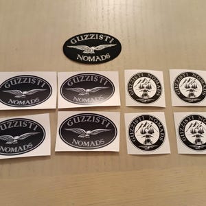 May include: A collection of black and white stickers featuring the text GUZZISTI NOMADS. Some stickers are oval-shaped with a flying eagle graphic, while others are circular with a mountain landscape and motorcycle silhouette design.