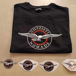 Może przedstawiać: Czarny T-shirt z białym logo orła i tekstem "Guzzisti Nomads". Na zdjęciu widoczne są również cztery białe naklejki z tym samym logo.