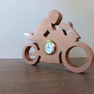 Könnte beinhalten: Eine holzgeschnitzte Uhr in Form eines Motorrads mit Fahrer-Silhouette. Das Zifferblatt ist goldfarben mit schwarzen Ziffern und Zeigern. Das Holz hat eine natürliche, hellbraune Oberfläche.