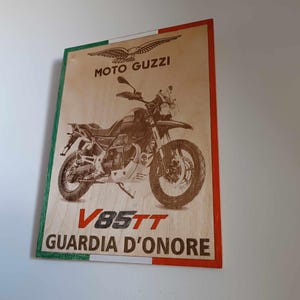 May include: Wooden sign with a Moto Guzzi V85TT motorcycle illustration. The sign has an Italian flag border and text reading "MOTO GUZZI" and "GUARDIA D'ONORE". A decorative piece for motorcycle enthusiasts.