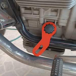 May include: An orange motorcycle wrench with a black gear-shaped head is positioned near the engine. The wrench is angled, with a loop at the end. The engine is silver and has the text "MOTO GUZZI" on it.