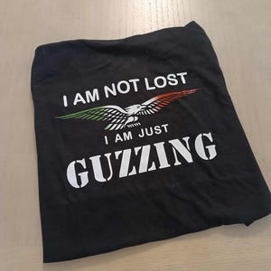 Può includere: T-shirt nero con la scritta "I AM NOT LOST I AM JUST GUZZING". La maglietta presenta una grafica di un'aquila bianca con accenti verdi, bianchi e rossi sulle ali. La maglietta è piegata su una superficie chiara.