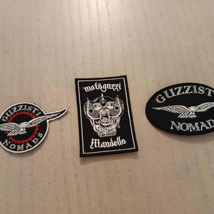 Może przedstawiać: Trzy haftowane naszywki z motywami Guzzi. Jedna jest okrągła z czerwono-czarnym wzorem, druga prostokątna z grafiką czaszki, a trzecia owalna z orłem. Naszywki na odzież.