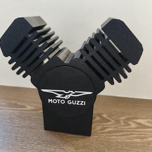 May include: A black V-twin engine model featuring prominent cooling fins and a central circular emblem. The emblem displays a white eagle graphic above the text MOTO GUZZI. The object has a matte finish and a distinct V-shaped configuration.