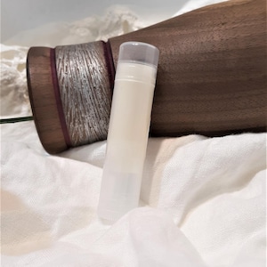 Puede incluir: Un tubo transparente de b&aacute;lsamo labial con un b&aacute;lsamo blanco cremoso en su interior. El tubo descansa sobre una tela blanca texturizada. Un objeto de madera oscura con detalles plateados se encuentra en el fondo.