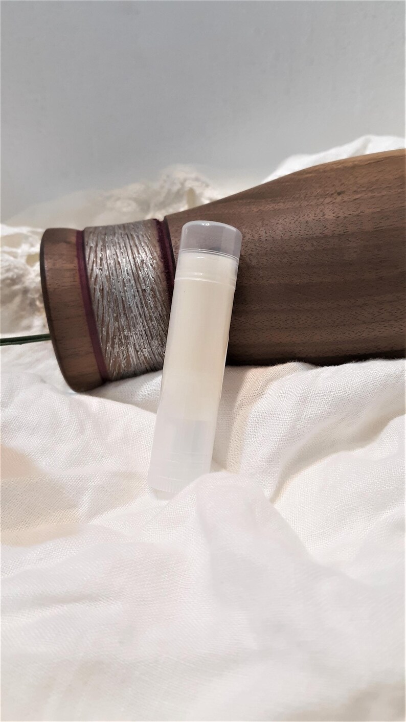 Puede incluir: Un tubo transparente de b&aacute;lsamo labial con un b&aacute;lsamo blanco cremoso en su interior. El tubo descansa sobre una tela blanca texturizada. Un objeto de madera oscura con detalles plateados se encuentra en el fondo.