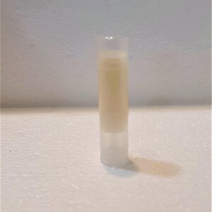 Puede incluir: Un tubo cil&iacute;ndrico transparente de b&aacute;lsamo labial lleno de un b&aacute;lsamo beige claro. El tubo est&aacute; de pie sobre una superficie blanca texturizada, proyectando una sombra. El fondo es de color claro y texturizado.