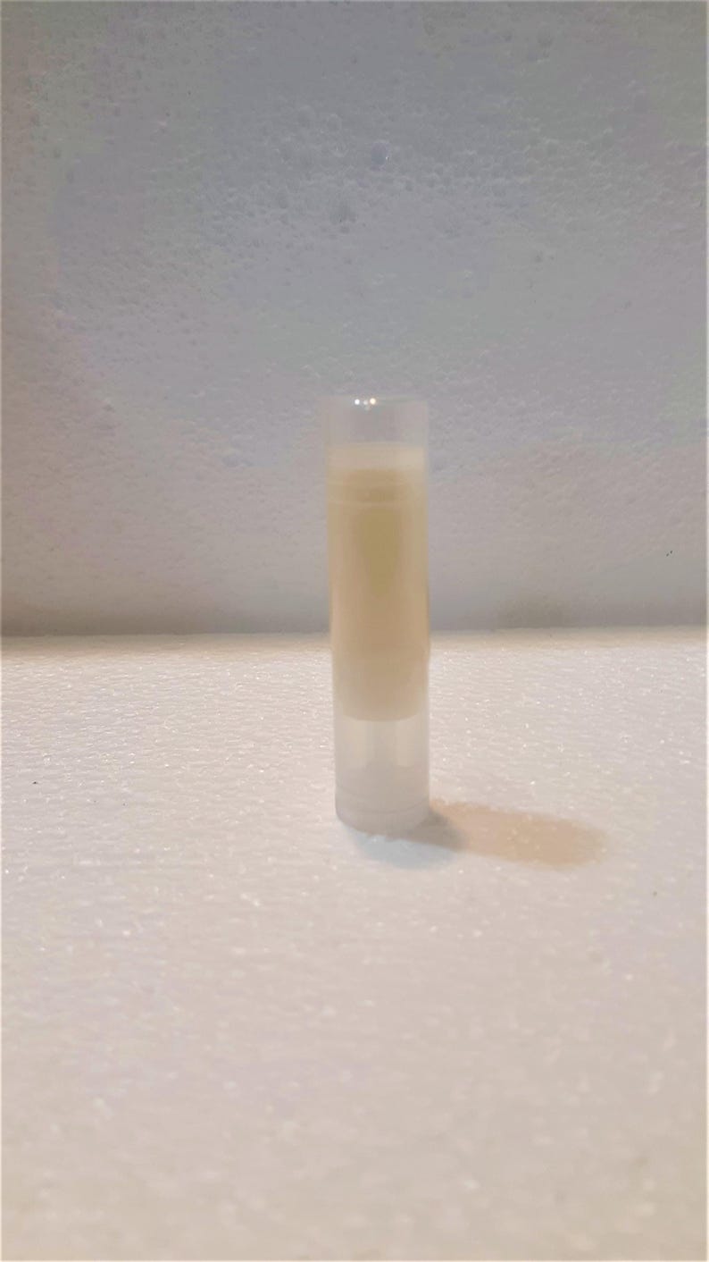 Puede incluir: Un tubo cil&iacute;ndrico transparente de b&aacute;lsamo labial lleno de un b&aacute;lsamo beige claro. El tubo est&aacute; de pie sobre una superficie blanca texturizada, proyectando una sombra. El fondo es de color claro y texturizado.