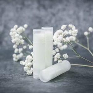 Puede incluir: Tres tubos de b&aacute;lsamo labial blancos y cil&iacute;ndricos, uno en posici&oacute;n horizontal, est&aacute;n dispuestos con ramitas de flores blancas de paniculata sobre un fondo gris texturizado. Los tubos son opacos y parecen estar llenos de una sustancia cremosa.