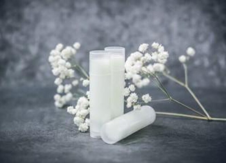Puede incluir: Tres tubos de b&aacute;lsamo labial blancos y cil&iacute;ndricos, uno en posici&oacute;n horizontal, est&aacute;n dispuestos con ramitas de flores blancas de paniculata sobre un fondo gris texturizado. Los tubos son opacos y parecen estar llenos de una sustancia cremosa.