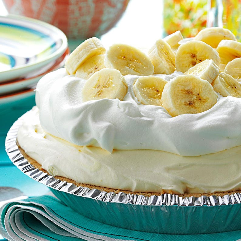 Puede incluir: Una tarta de crema de pl&aacute;tano en una bandeja de aluminio, cubierta con crema batida y rodajas de pl&aacute;tano. La tarta tiene una corteza dorada y un relleno cremoso. El postre est&aacute; sobre una superficie azul con una servilleta turquesa.