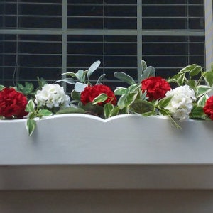 May include: A white window box planter with red and white flowers. The planter is mounted on a white wall.