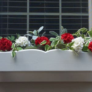 May include: A white window box planter with red and white flowers. The planter has a scalloped edge and is filled with green foliage.