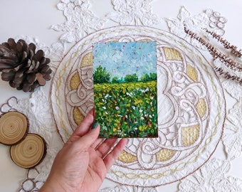 May include: A small, rectangular oil painting depicts a vibrant landscape. The artwork showcases a field of yellow and white flowers under a blue sky with green trees in the distance. The painting is held against a white, embroidered background.