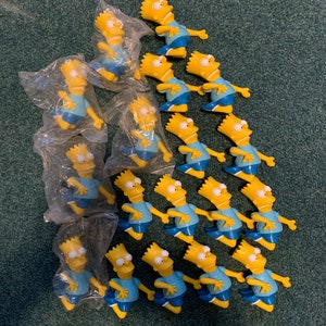 May include: A collection of 18 yellow plastic figurines of Bart Simpson from The Simpsons. Each figurine is wearing a blue shirt and blue shorts and is packaged in a clear plastic bag.