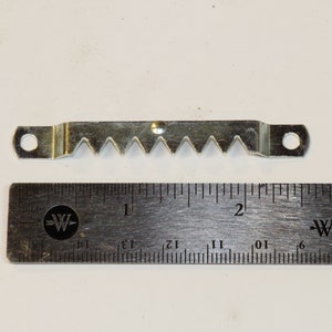 May include: A silver-toned sawtooth picture hanging hook with two holes. The hook is 5 cm long and is measured against a ruler.