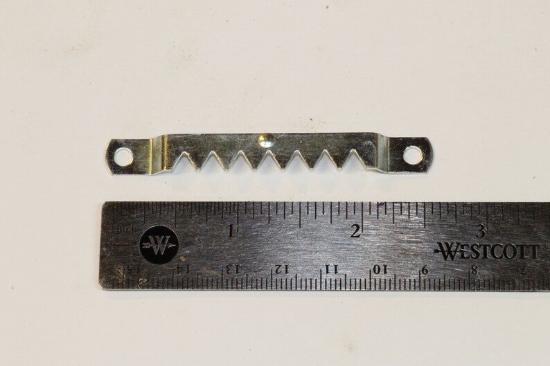 May include: A silver-toned sawtooth picture hanging hook with two holes. The hook is 5 cm long and is measured against a ruler.