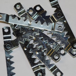 May include: A close-up of a pile of silver metal picture hanging hooks with sawtooth edges. The hooks have a small hole at the top for hanging.
