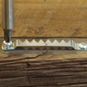 May include: A close-up of a silver picture hanging hook being screwed into a piece of wood with a screwdriver.