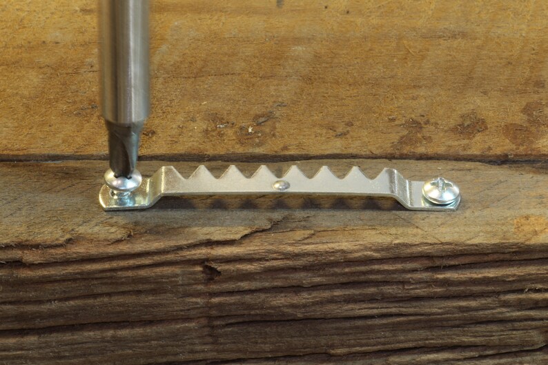 May include: A close-up of a silver picture hanging hook being screwed into a piece of wood with a screwdriver.