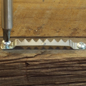 May include: A close-up of a metal sawtooth picture hanger being screwed into a piece of wood. The hanger is silver and has a series of triangular teeth. A screwdriver is being used to secure the screw.
