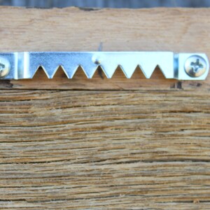May include: A silver metal sawtooth picture hanging hook with two screws. The hook is attached to a piece of wood.