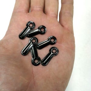 May include: A close-up of six black metal eyelet rings. The rings are small and have a flat, rectangular shape with a round hole in the center.