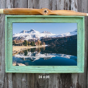 May include: A framed landscape print featuring a mountain range reflected in a lake, with a light green wooden frame. A wooden propeller is above the frame. The print measures 24 x 36 inches.