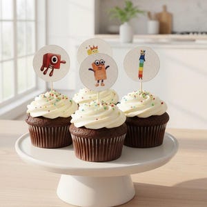 May include: Four chocolate cupcakes with white frosting and colourful sprinkles on a white cake stand. Each cupcake has a round topper with a cartoon character. The cupcakes are in front of a bright kitchen.