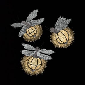 May include: Three embroidered fireflies on a black background. Each firefly has gray wings, a gray body, and a glowing yellow orb for a tail. The orbs are surrounded by a spiky, golden-brown halo.