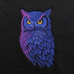 May include: An embroidered owl design on a black fabric background. The owl features intricate stitching in shades of blue and purple, with bright yellow eyes. The embroidery creates a textured, layered appearance of feathers across the owl's body.