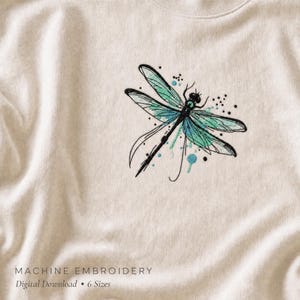May include: A cream-colored sweatshirt featuring an embroidered dragonfly design. The dragonfly has black outlines with teal and light blue wing details, accented by small black dots and blue paint-like splatters. Text reads MACHINE EMBROIDERY.