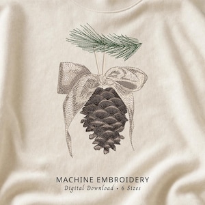 Peut inclure: Un sweat-shirt couleur crème avec une broderie représentant une pomme de pin brune suspendue à une branche de pin verte par un ruban clair. Le texte sous la broderie indique MACHINE EMBROIDERY Digital Download 6 Sizes.