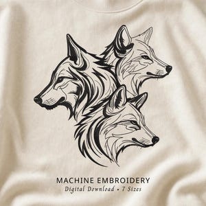 May include: A cream-colored sweatshirt featuring a black machine embroidery design of three wolf heads arranged in a cluster. Below the wolves, the text reads MACHINE EMBROIDERY Digital Download 7 Sizes in a clean, black serif font.