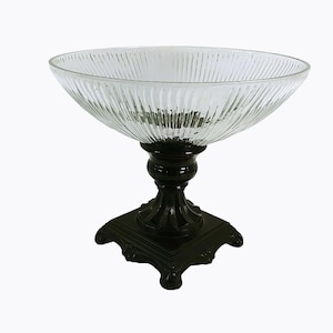 May include: A clear glass bowl with a fluted design sits on a black pedestal with a square base. The pedestal has ornate details and a dark wood finish.