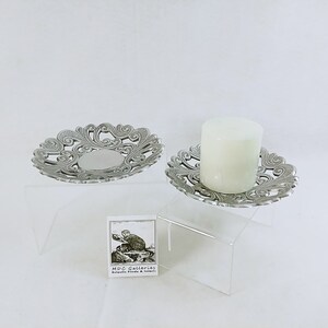May include: Two silver candle holders with ornate designs. The candle holders are on clear acrylic stands. The candle holder on the right has a white candle inside.