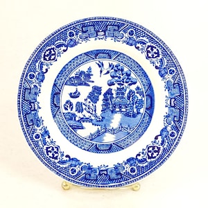 May include: A blue and white porcelain plate with a willow pattern design. The plate has a white background with a blue border and a central design of a willow tree, a bridge, and a pagoda.