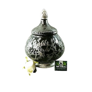 May include: A decorative ceramic jar with a silver floral design on a dark green background. The jar has a lid with a white finial. The jar is sitting on a white base.
