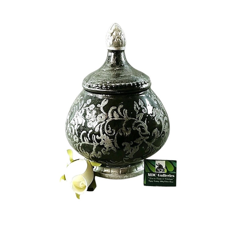 May include: A decorative ceramic jar with a silver floral design on a dark green background. The jar has a lid with a white finial. The jar is sitting on a white base.