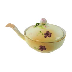 May include: A small, ceramic serving dish with a lid. The dish is pale yellow with purple floral accents and a pink tulip-shaped handle on the lid. It has a long, curved handle on the side. The rim is trimmed with gold.