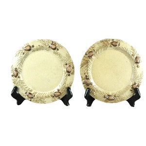 May include: Two decorative ceramic plates with a raised design of pine branches and cones. The plates are a cream color with brown accents and are displayed on black stands.