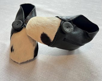 May include: A pair of black leather slippers with a white and black cowhide pattern. The slippers have a button closure.