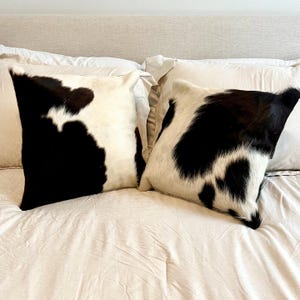 May include: Two square throw pillows with a cowhide pattern. The pillows feature a natural white background with irregular black patches. They are placed on a bed with white bedding, creating a rustic and stylish aesthetic.