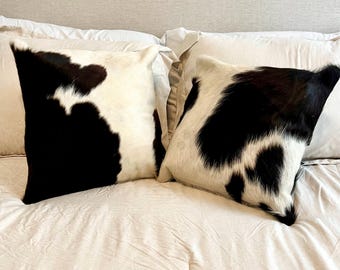 May include: Two square throw pillows with a cowhide pattern. The pillows feature a natural white background with large black patches. They are placed on a bed with white bedding.
