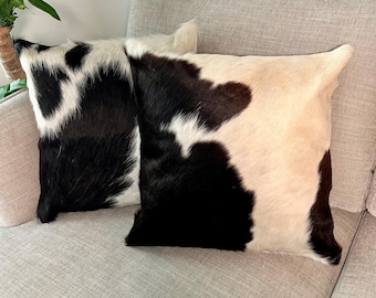 May include: Two black and white cowhide throw pillows with a soft, plush texture. The pillows are arranged on a light gray couch.
