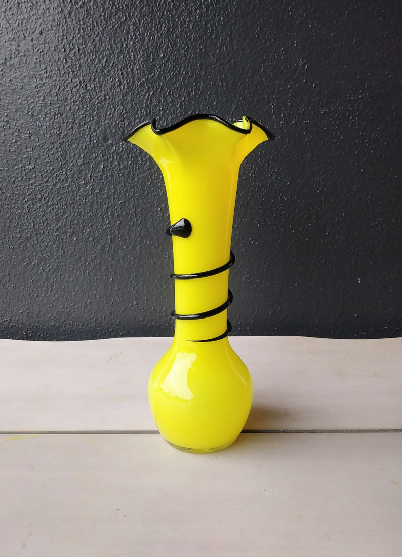 May include: A yellow glass vase with a black swirl design. The vase has a wide mouth and a narrow base.