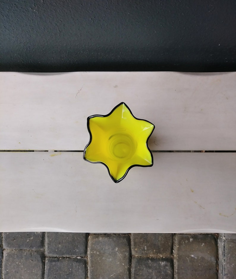 May include: A yellow glass vase with a star-shaped opening and black trim.