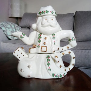 May include: White ceramic teapot shaped like Santa Claus. The teapot is decorated with green holly leaves and red berries. The teapot has a gold handle and spout.